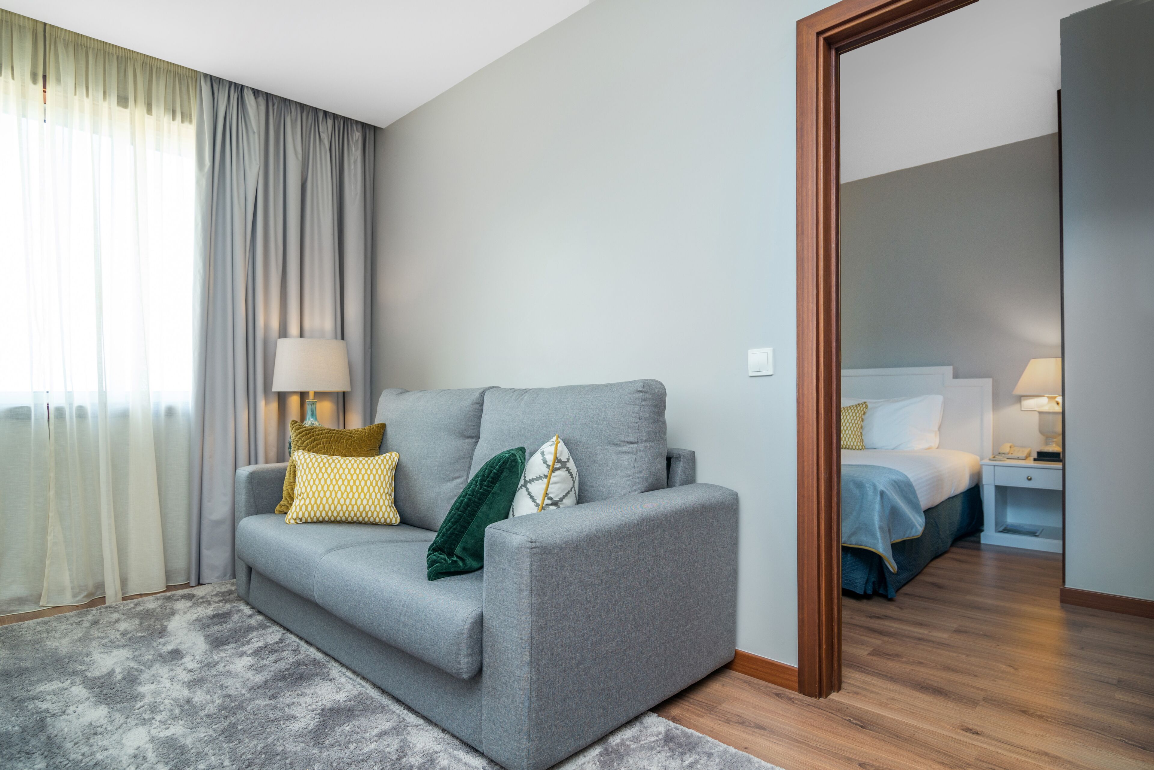 A modern living room with a grey sofa, a wooden floor, and a large mirror reflecting the room.