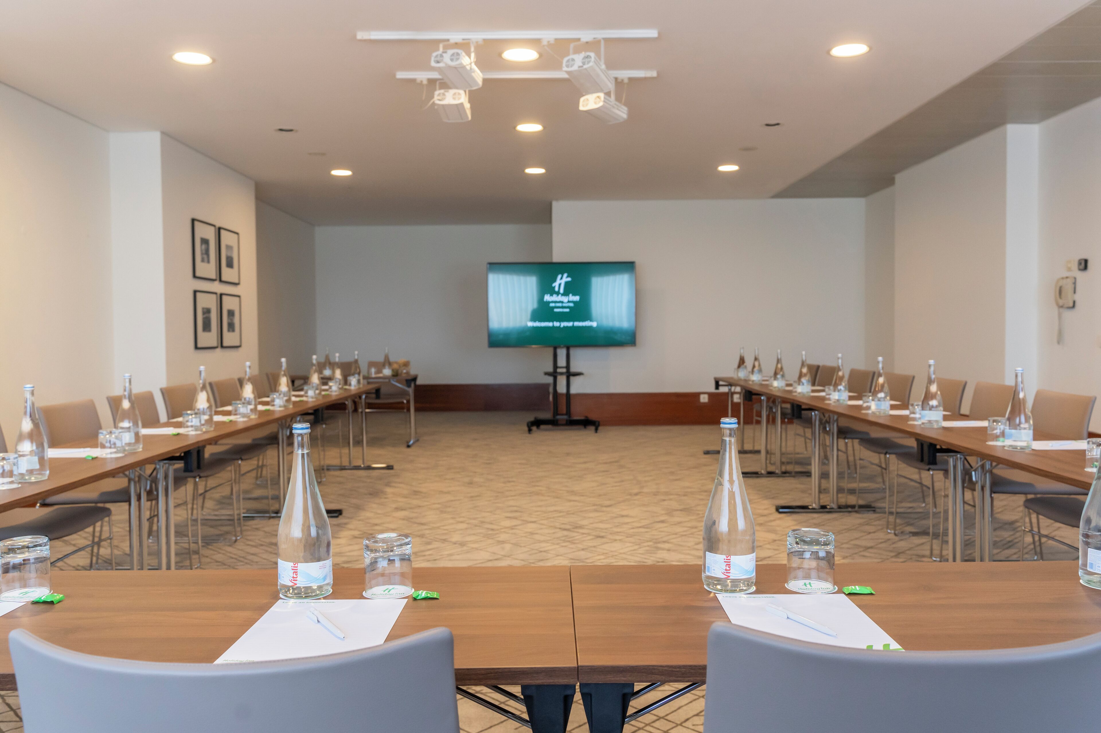 Een vergaderzaal met lange tafels, stoelen, waterflesjes en een scherm waarop een welkomstboodschap staat.