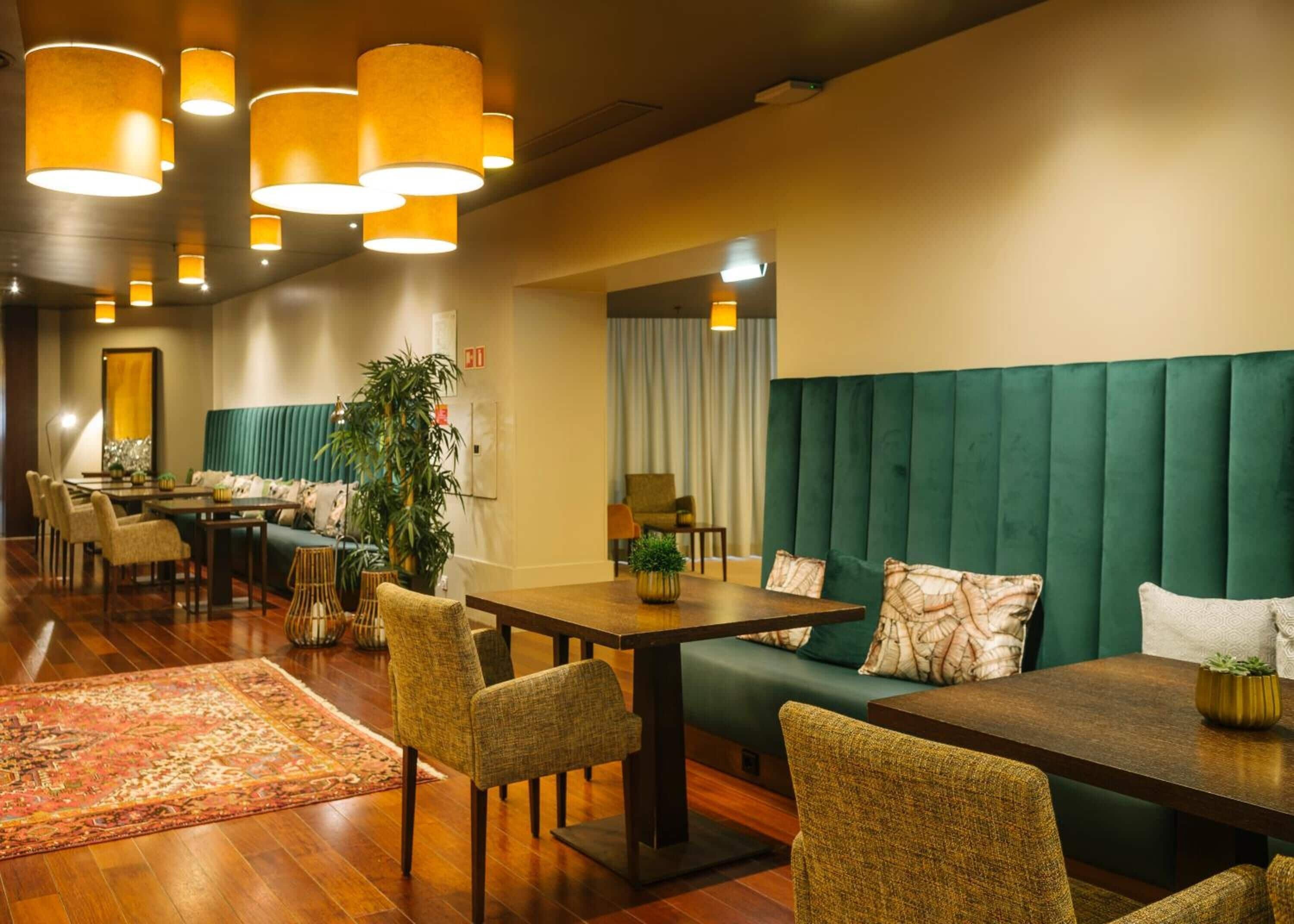 A hotel lobby with a green upholstered booth, wooden tables, and chairs. There are hanging lamps and a large potted plant.