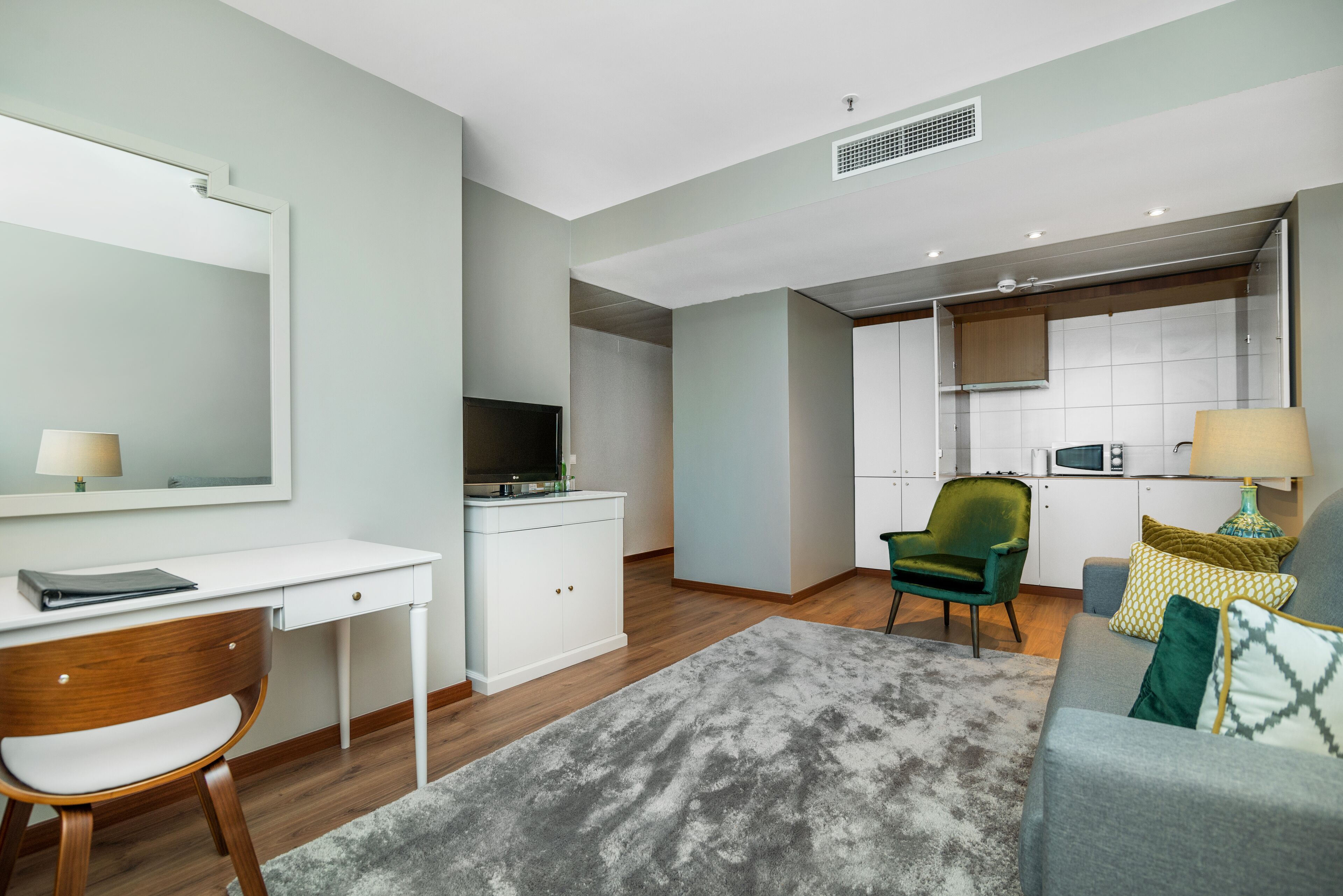 A modern living room with a grey sofa, a green armchair, a white desk, and a kitchen area in the background.