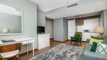 Un salon moderne avec un canapé gris, un fauteuil vert, un bureau blanc et un coin cuisine à l’arrière-plan.