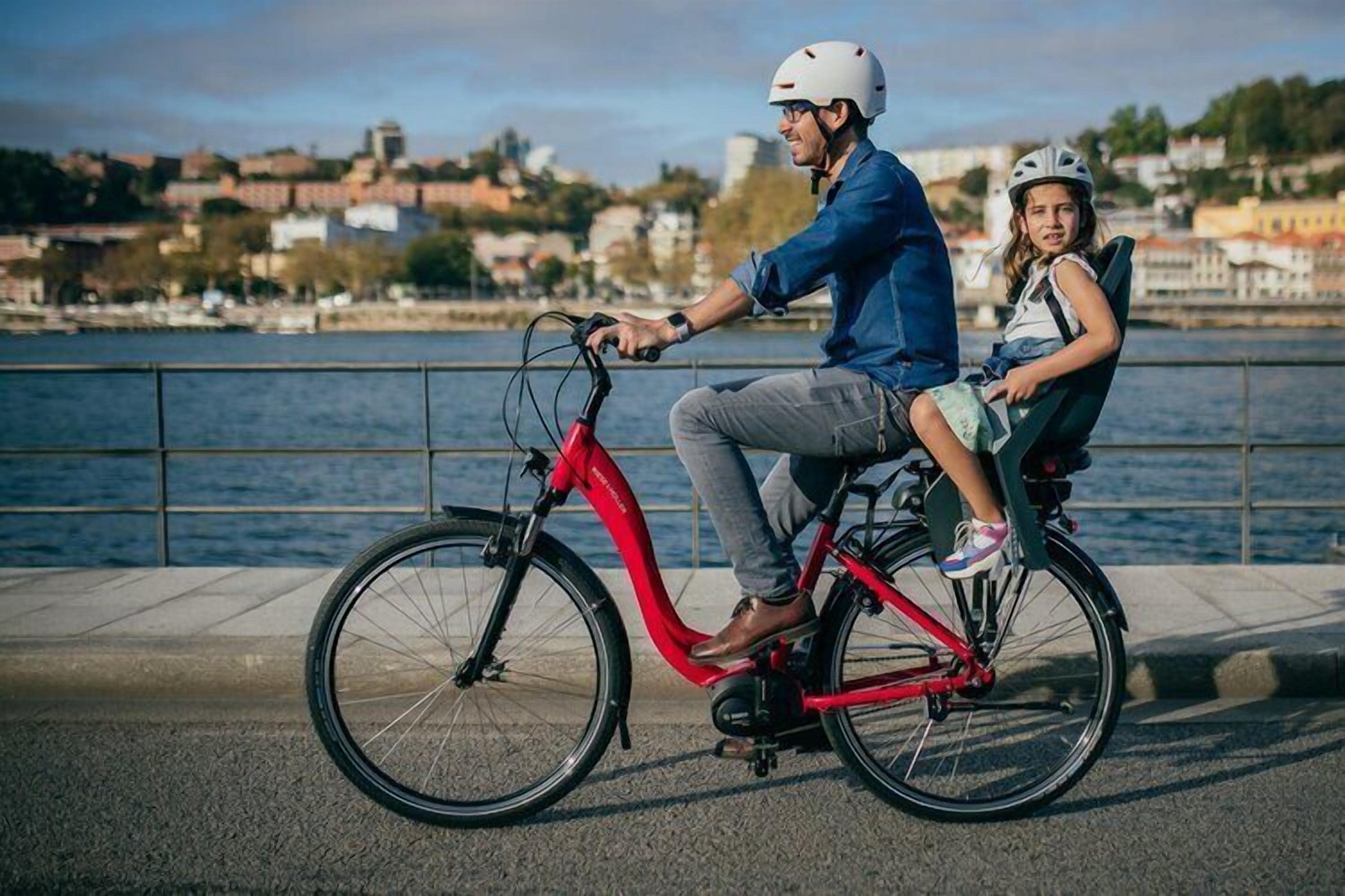 Un vélo électrique rouge, sur lequel sont assis un enfant et un adulte, est garé au bord de l’eau, avec des bâtiments en arrière-plan.