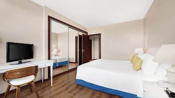 A hotel room with a bed, a desk with a TV, a chair, a bedside table with a lamp, and a wardrobe.