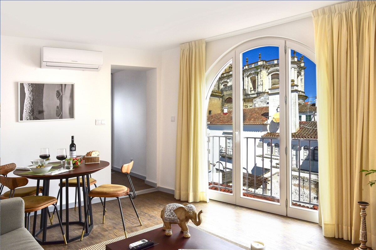 A modern living room with a dining area, a large window with a view of historic buildings, and a small elephant figurine on the floor.