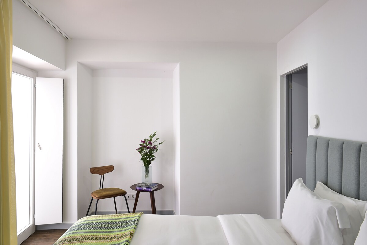 A hotel room with a bed, a chair, a small table with a vase of flowers, and a door.
