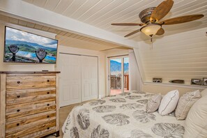Un dormitorio con cama, cómoda de madera, ventilador de techo, televisor y vista a montañas a través de una puerta.