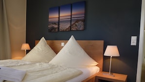 A neatly made bed with white linens and pillows, a wooden headboard, a bedside table with a lamp, and a wall-mounted artwork depicting a sunset over a beach. - Baltic Sea Lodges, Deck 1, elevator, wellness area, close to the beach, family friendly (Niendorf)