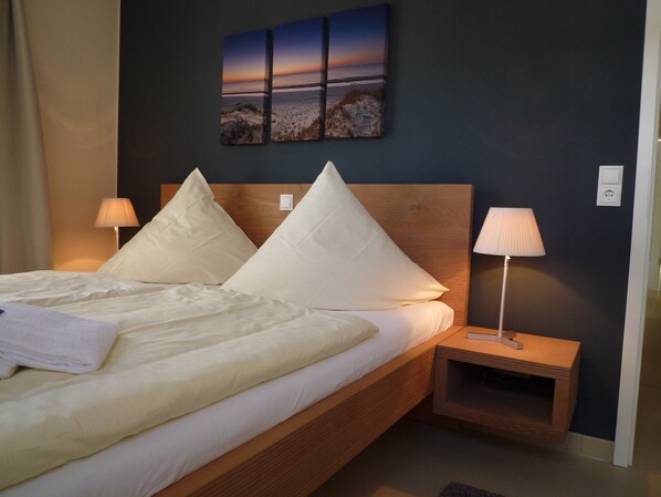 Una cama bien tendida con sábanas y almohadas blancas, cabecera de madera, mesita de noche con lámpara y una obra de arte en la pared que representa un atardecer sobre una playa.