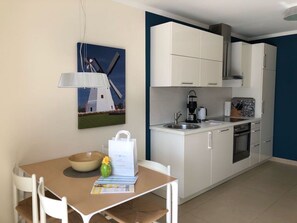 A compact kitchen with white cabinets, a sink, and a stove. There's a dining table with chairs and a bowl. A painting of a windmill is on the wall.