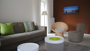 Un salon moderne avec un canapé gris, une table basse ronde, un fauteuil en osier et un fauteuil de couleur grise. Un tableau représentant un paysage est accroché au mur brun.