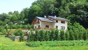 A two-story house with a balcony, surrounded by lush greenery and a wooden bridge. - B&B Il Mortirolo (Mazzo di Valtellina)