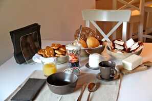 Une table mise pour le petit-déjeuner, avec divers aliments, notamment des viennoiseries, un verre de jus d’orange et un bol de yaourt agrémenté de garnitures.