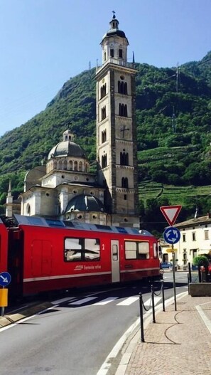 Un train rouge, portant l’inscription « Ferrovie dello Stato » sur le côté, passe devant un bâtiment historique doté d’un clocher et de coupoles, le tout se détachant sur un arrière-plan de collines verdoyantes.