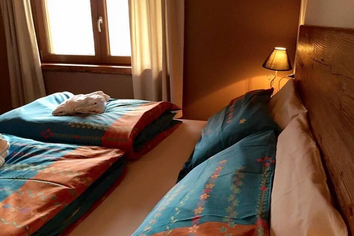 A hotel room with two beds, a window with curtains, and a bedside lamp.