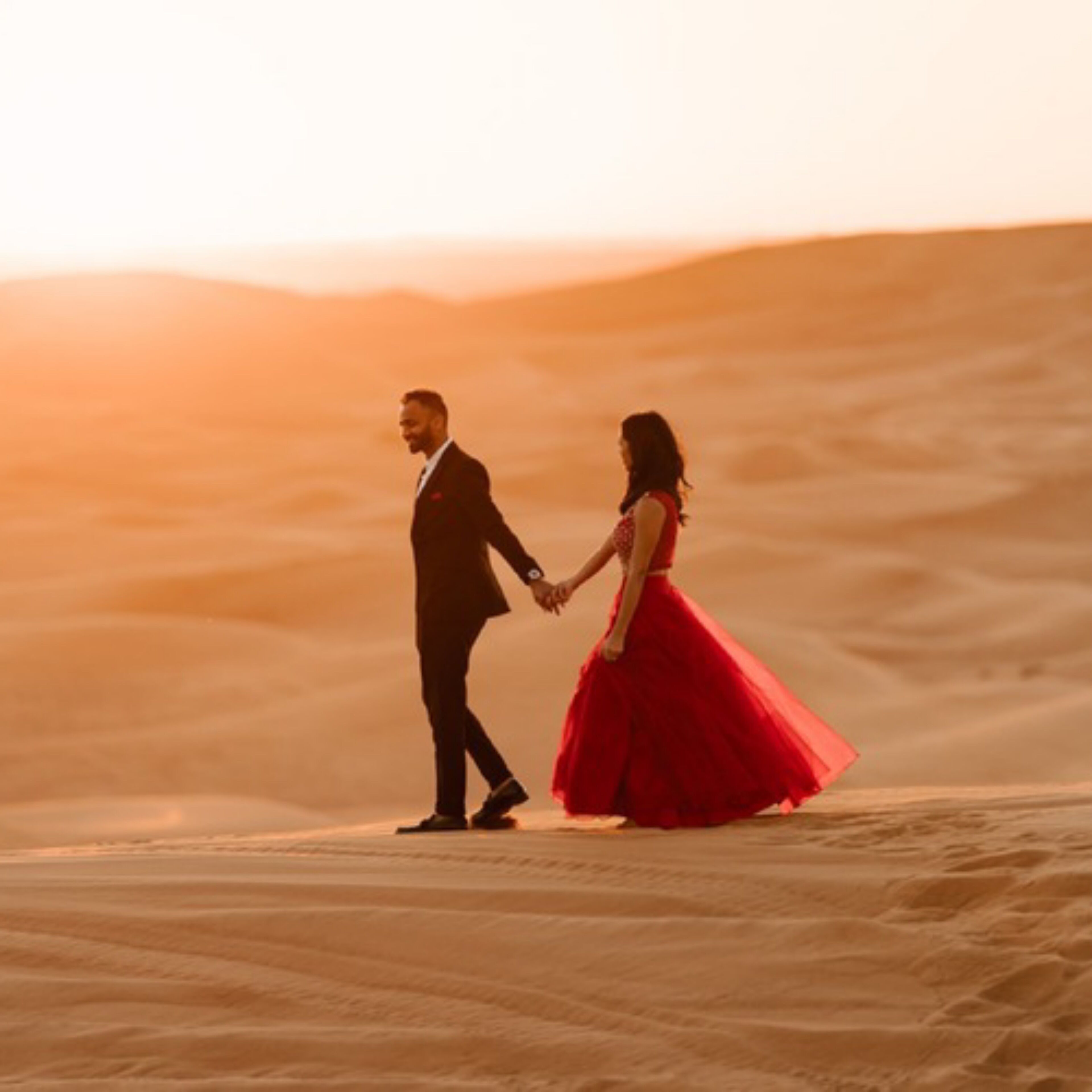 A couple holding hands on a sandy beach at sunset.
