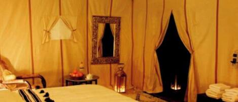 A tented room with a bed, a mirror, a table with a lamp, and a fireplace.