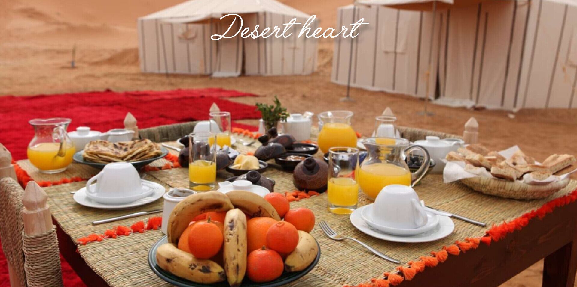Mesa al aire libre con temática desértica, ofreciendo diversos alimentos para el desayuno, como frutas, pan y bebidas.