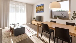 Een moderne woonkamer met een houten eettafel, zwarte stoelen, een grijze poef, een flatscreen-tv en een groot raam met uitzicht op bomen.