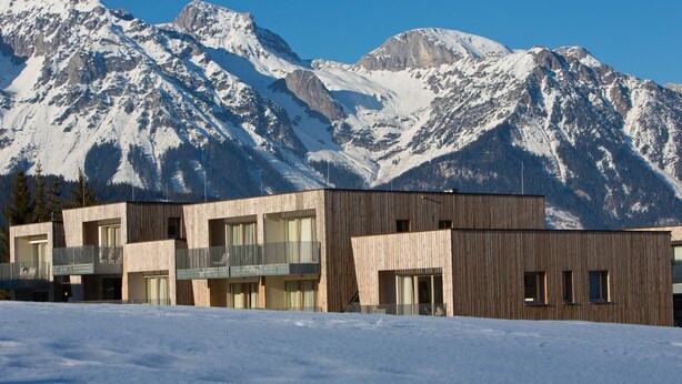 Een modern gebouw met een houten gevel, grote ramen en balkons, met een besneeuwd berglandschap op de achtergrond.