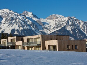 A modern building with a wooden facade, large windows, and balconies, set against a backdrop of snow-covered mountains.