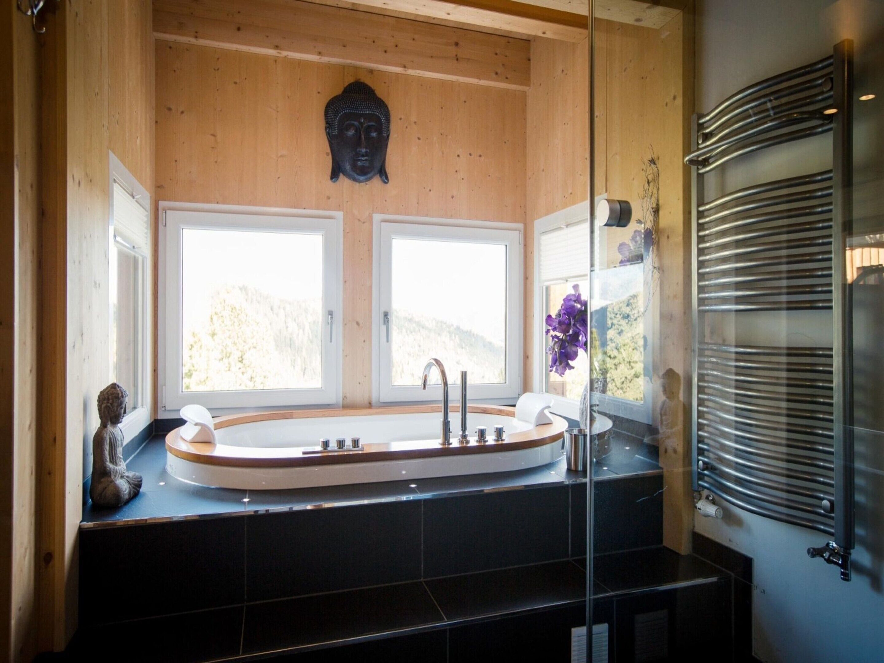 A modern bathroom with a freestanding bathtub, two windows, a Buddha statue, and a glass shower enclosure.