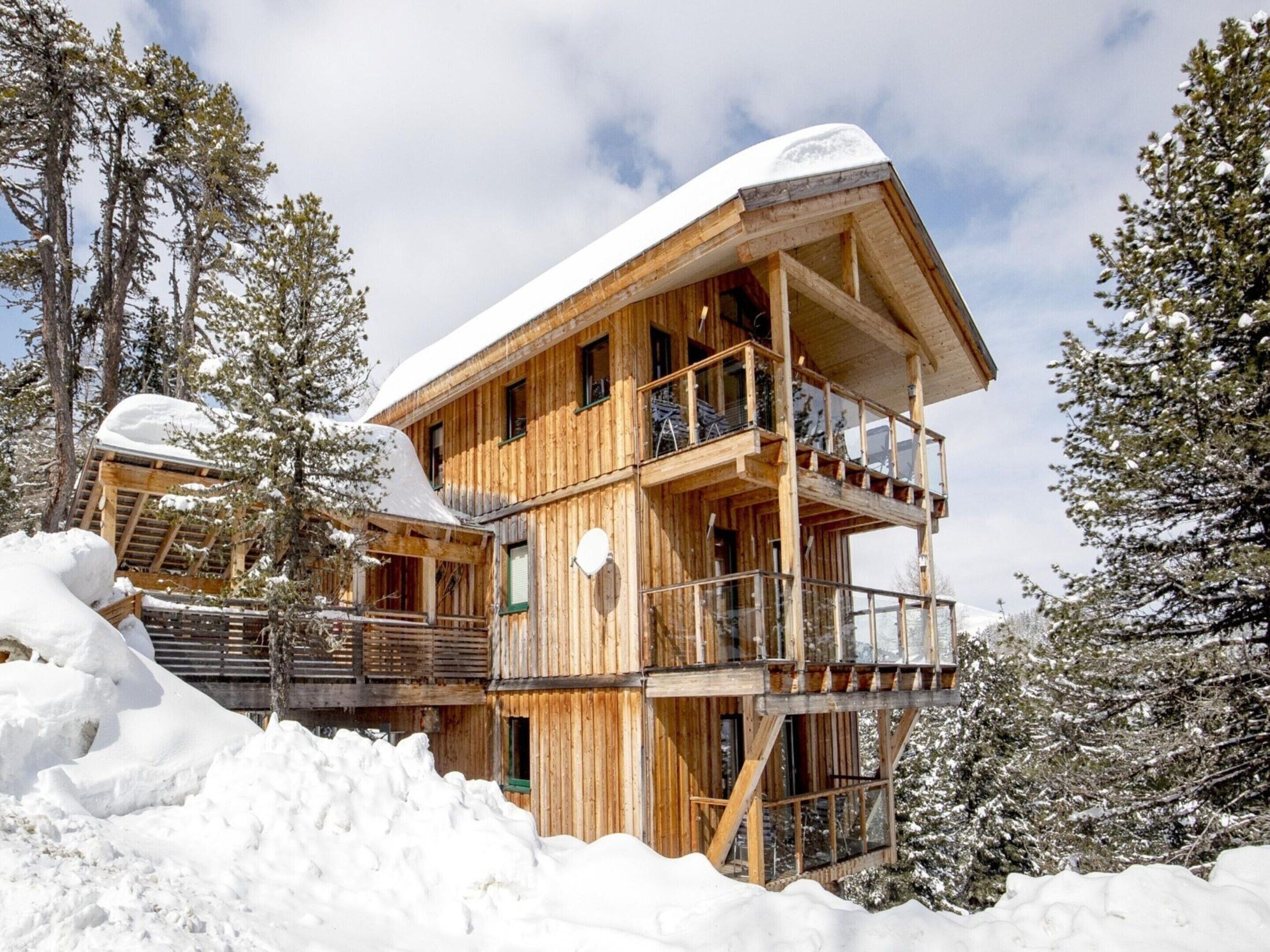 A wooden chalet with a balcony and a snow-covered roof, surrounded by trees.