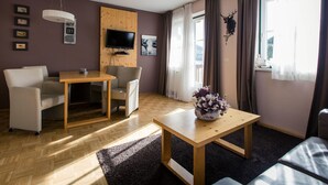 A modern living room with a wooden table, a sofa, a TV mounted on a wooden panel, and a window with curtains. - Cozy Apartment near Ski Area in Turracherhohe (Stadl-Predlitz)