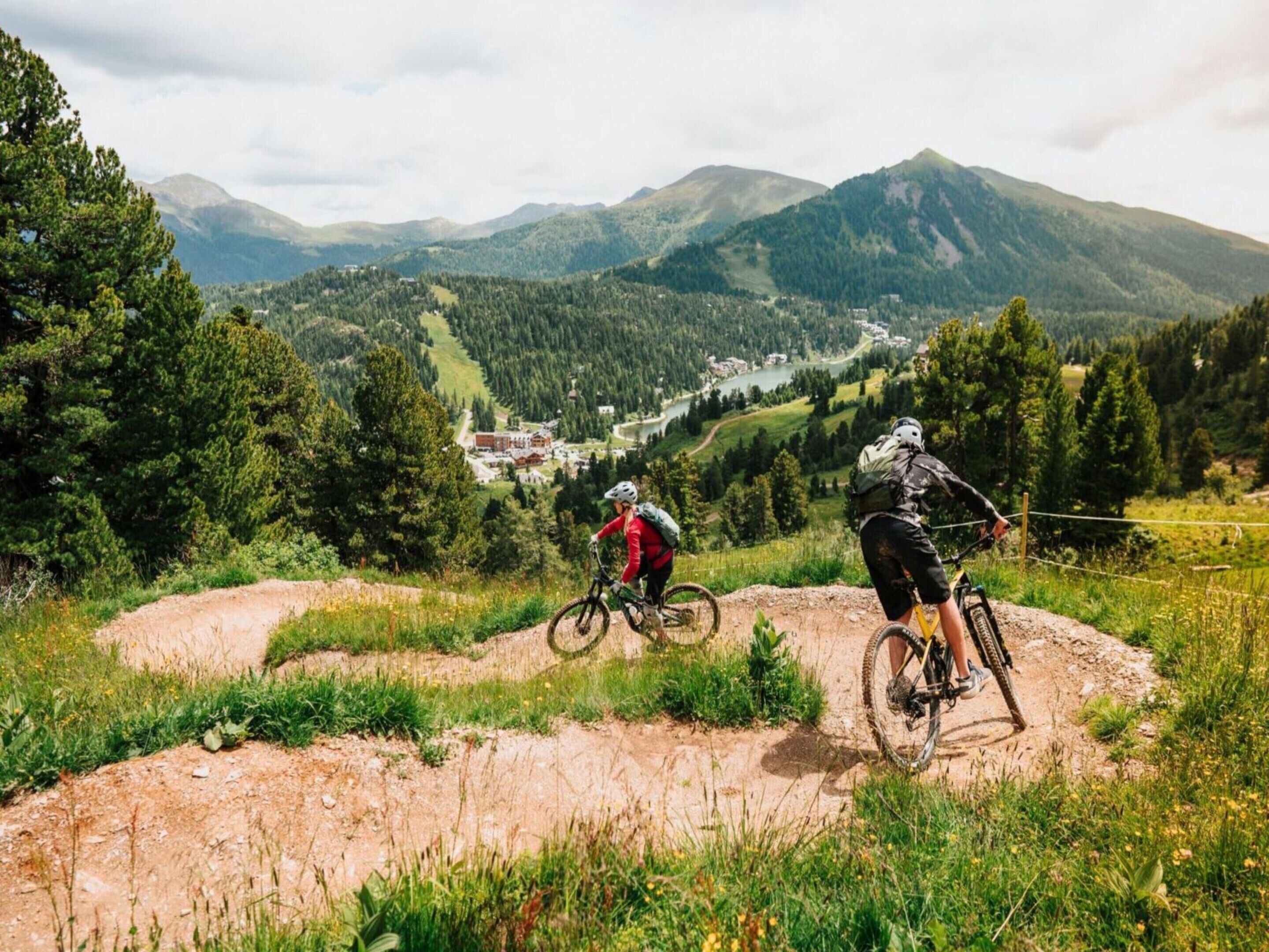 Dois ciclistas de montanha numa trilha de terra, com uma vista panorâmica de um vale e montanhas ao fundo.