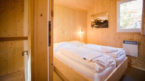 A wooden cabin bedroom with a bed, a wall-mounted light, and a window with a view of trees.