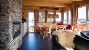 A cozy cabin interior with a stone fireplace, wooden dining area, and large windows offering a view of the outdoors.