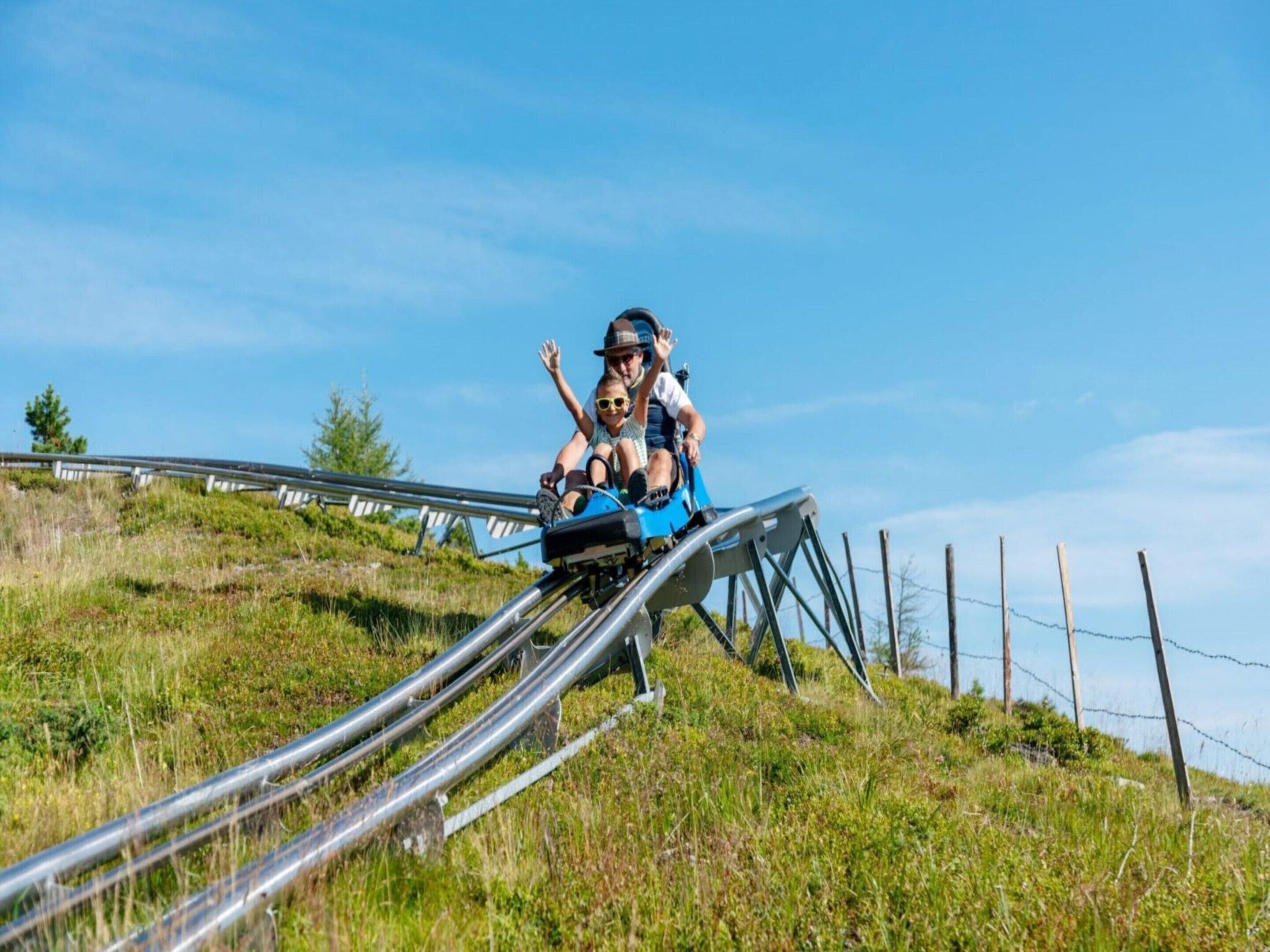 Une personne est assise sur un engin ressemblant à une luge, qui se déplace sur une piste métallique. En arrière-plan, on aperçoit une colline herbeuse et une clôture.