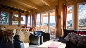 A cozy wooden cabin interior with a dining area, seating, and large windows offering mountain views.