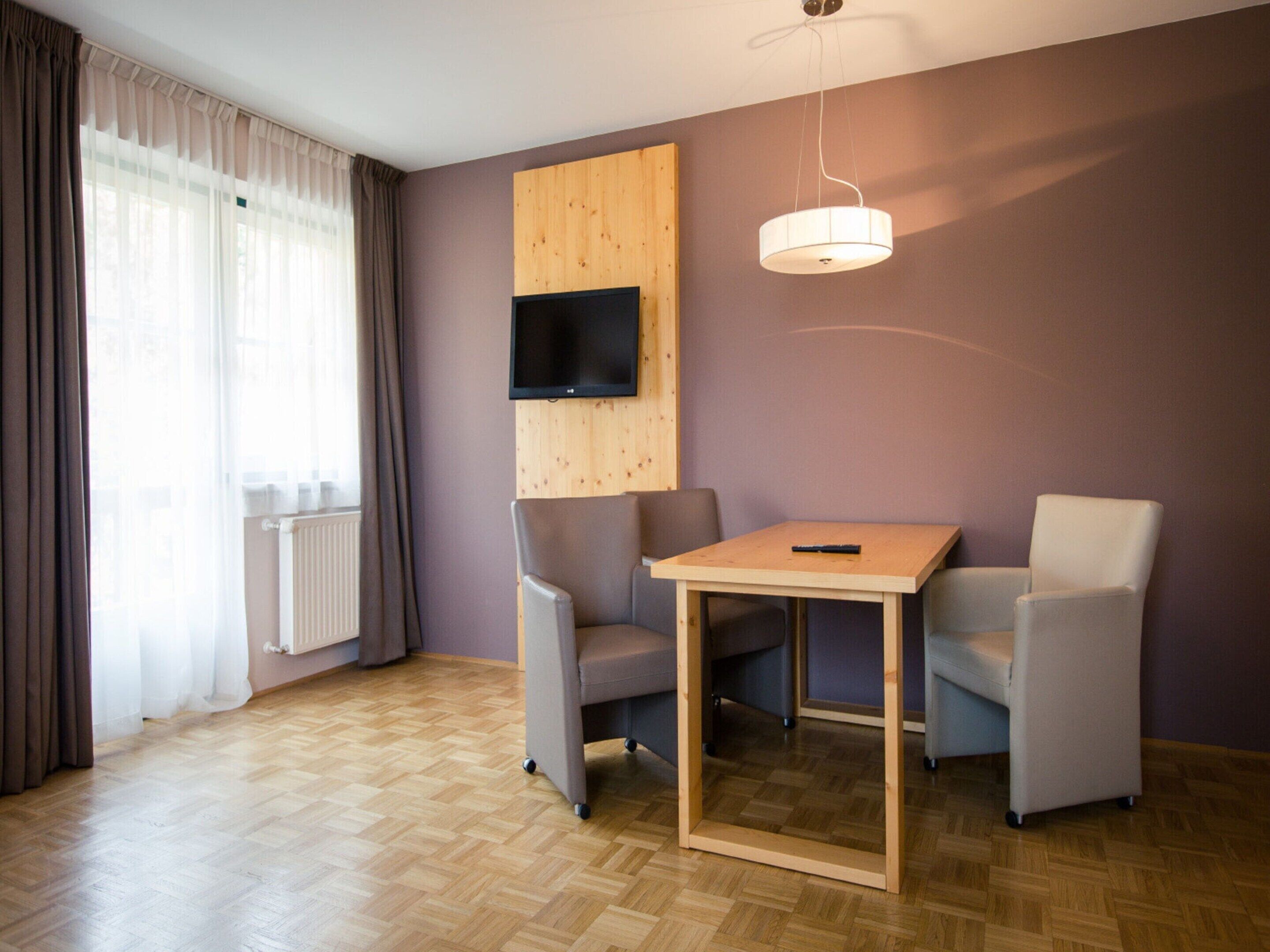 Una habitación moderna con suelo de madera, un televisor montado en un panel de madera, una mesa con dos sillas y una ventana con cortinas.
