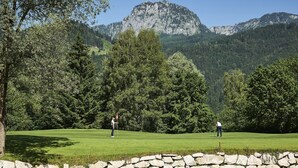 Un parcours de golf avec un étang et un mur de pierres, entouré d’arbres et de montagnes.
