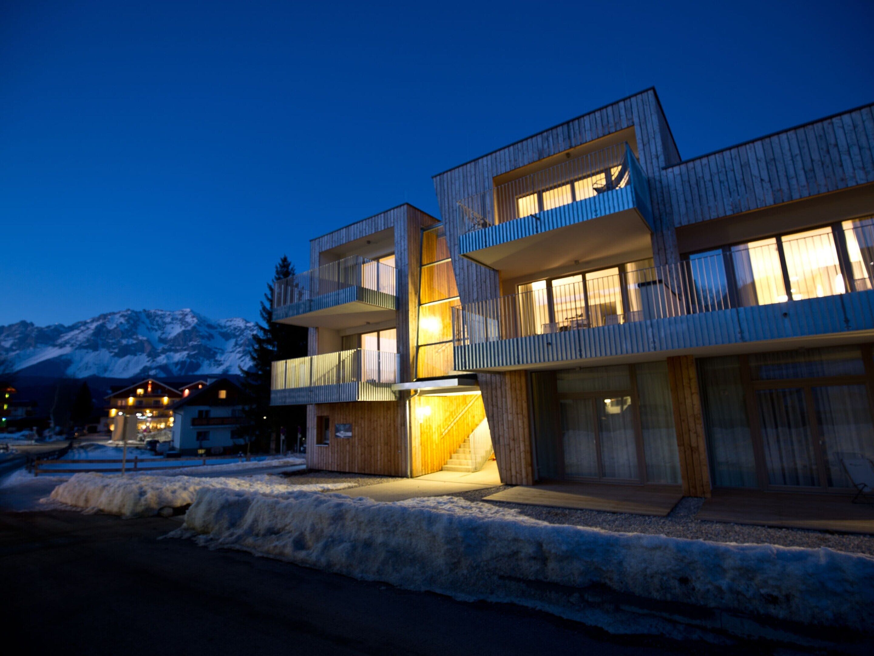 Un bâtiment moderne à plusieurs étages, doté de balcons et de grandes fenêtres, qui se dresse sur fond de montagnes enneigées.