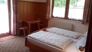 Een kleine, knusse kamer met een houten bed, een bureau en een stoel. Er is een raam met gordijnen, en een radiator is zichtbaar aan de muur.