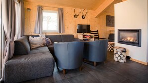 Ein gemütliches Wohnzimmer mit einer Couch, zwei Sesseln, einem Flachbildfernseher, einem Kamin und einem Fenster mit Vorhängen.