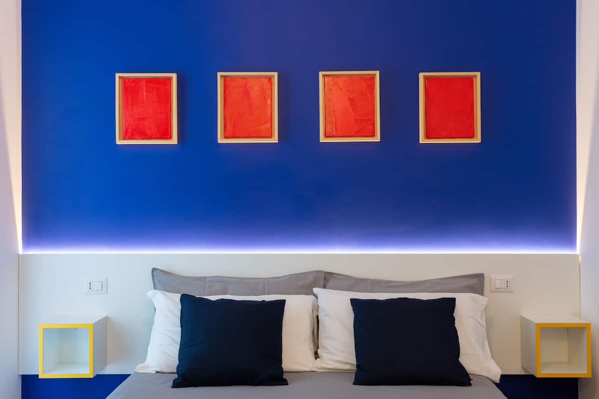 A modern bedroom with a blue accent wall, a bed with grey and white bedding, and four red-framed paintings.