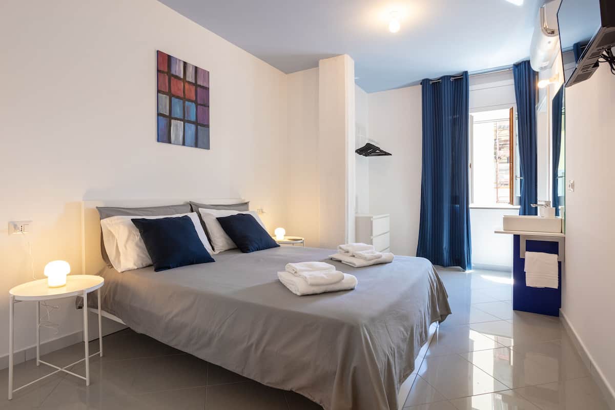 A modern hotel room with a neatly made bed, a bedside table with a lamp, a bathroom with a mirror, and a view of the outside.