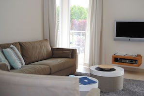 A modern living room with a brown sofa, a flat-screen TV mounted on the wall, a small wooden side table, and a round ottoman. - Baltic Sea lodges, apartment deck 3, wellness with sauna, fitness, elevator (Niendorf)