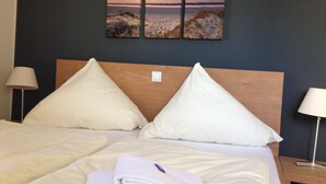 Quarto de hotel com cama, duas almofadas, toalha dobrada e uma obra de arte na parede retratando uma cena de praia.