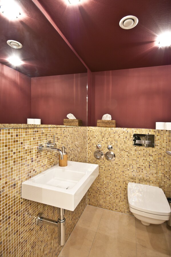 A bathroom with a tiled mosaic wall, a white sink with a wooden soap holder, a toilet, and a tissue box on the wall.