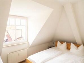 Un dormitorio con techo inclinado, una ventana, un radiador, una cama con almohadas y una mesita de noche.