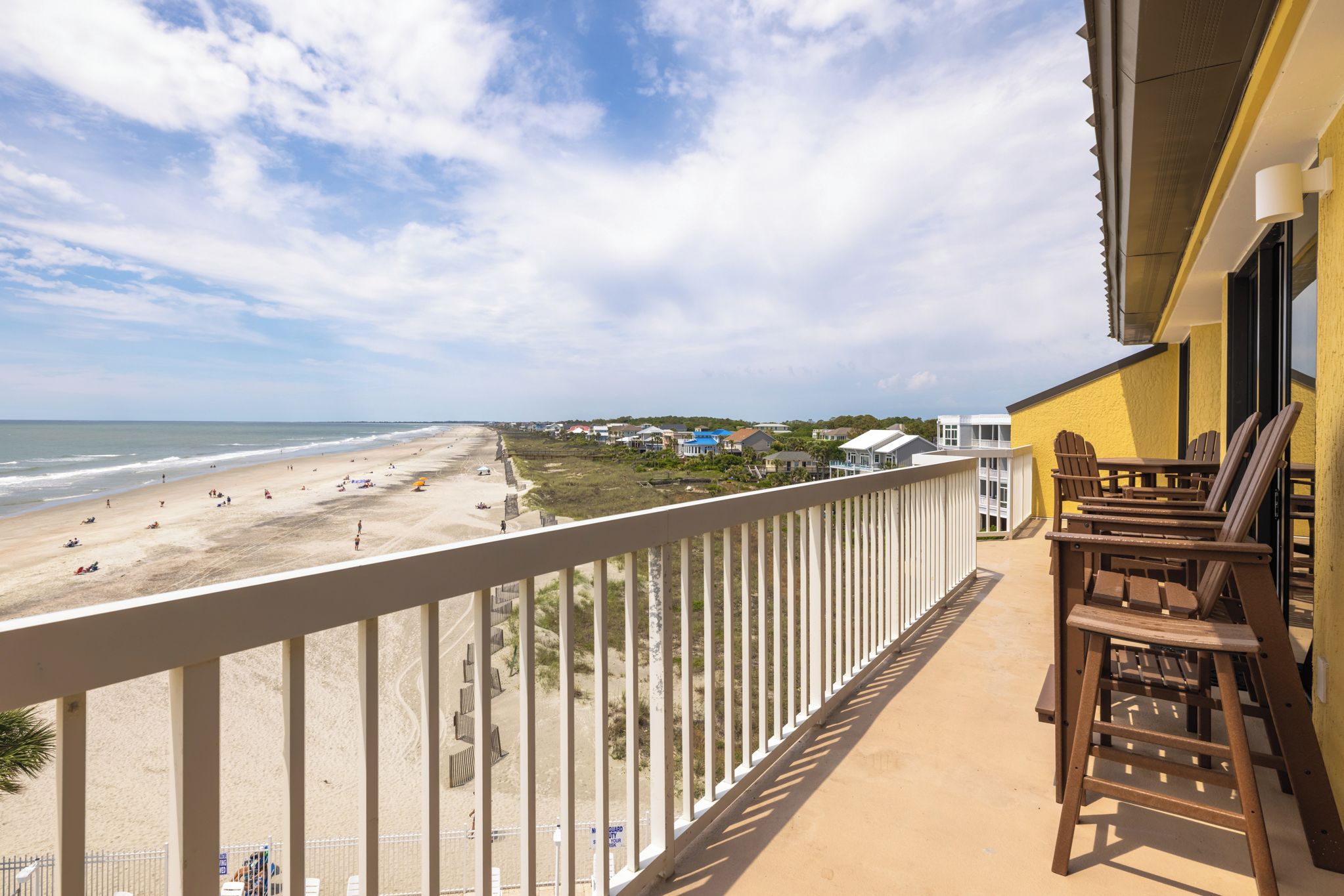 Een balkon met uitzicht op een strand en de oceaan, houten stoelen en een leuning.