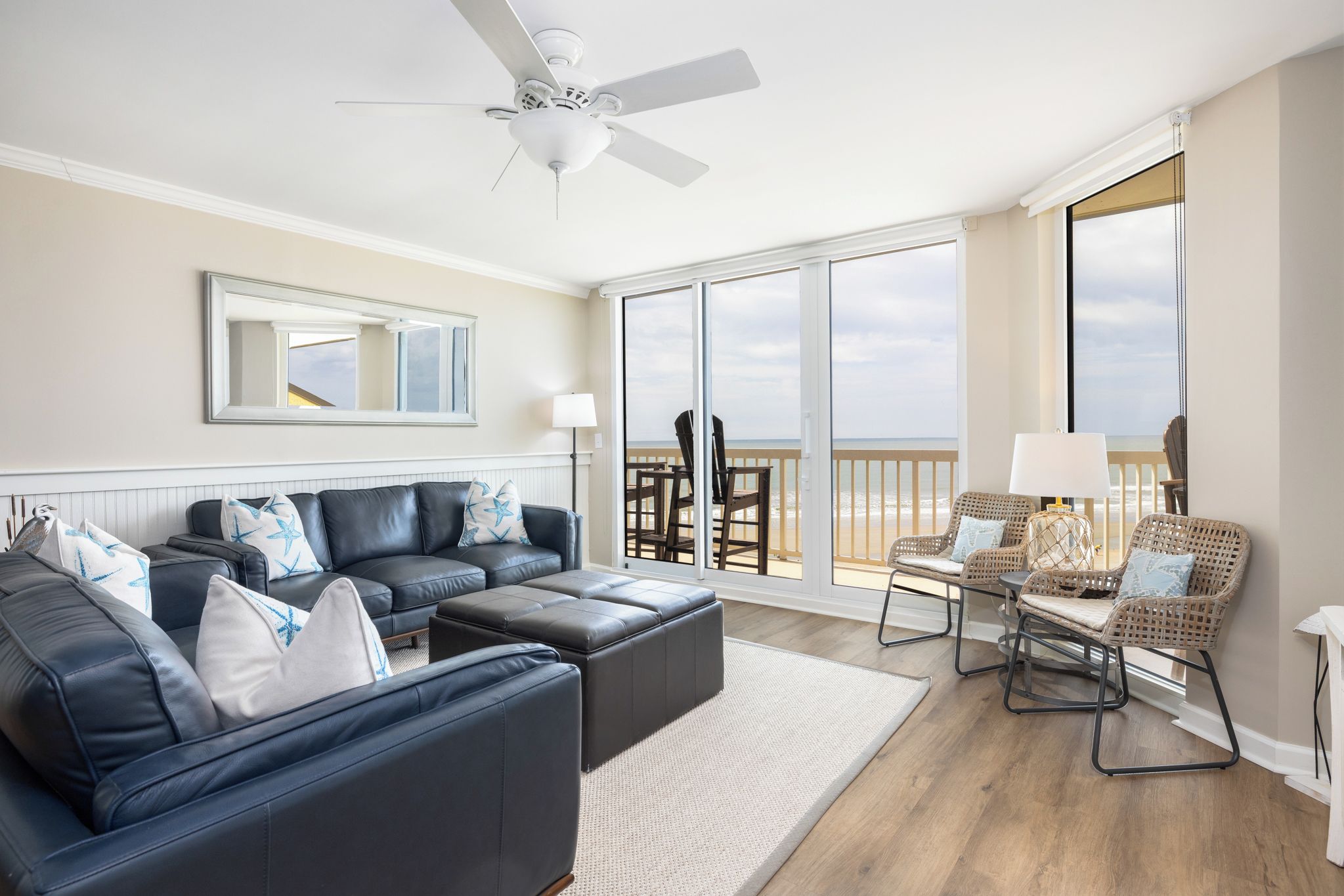 Un salon comprenant un grand canapé, une table basse et un coin repas avec des chaises en osier. Il y a également un balcon avec vue sur la plage.