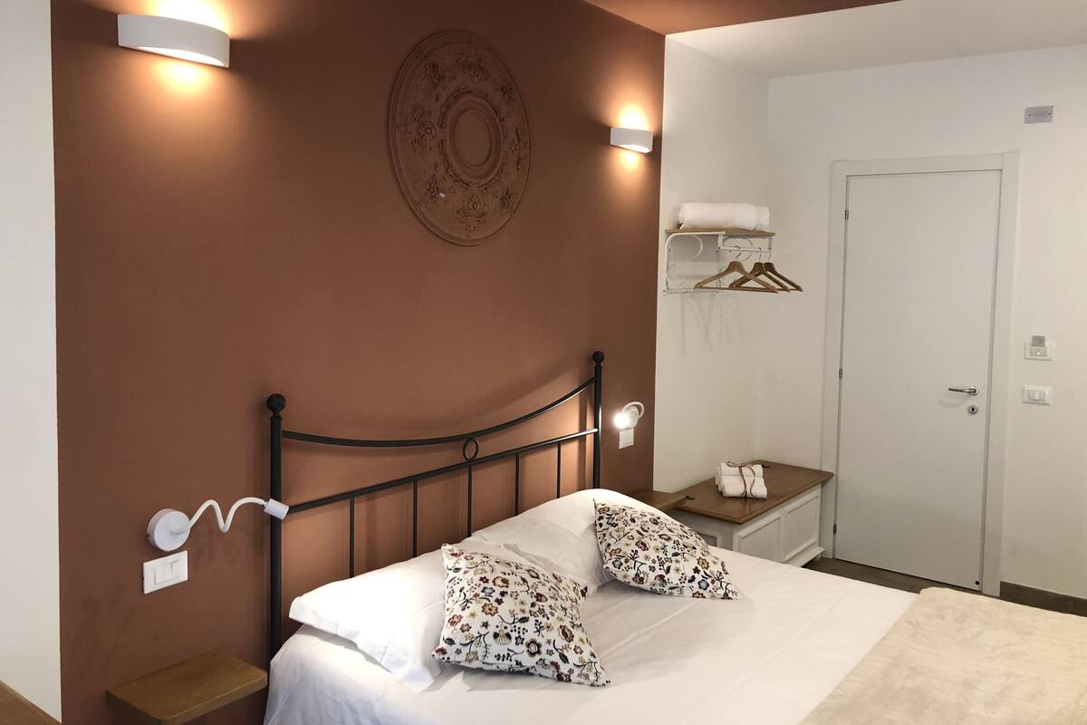 A bedroom with a bed, two wall-mounted lights, a bedside table with a towel, a coat rack, and a door.