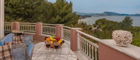 A balcony with wicker furniture, a table with fruit and wine glasses, and a view of a body of water and hills.