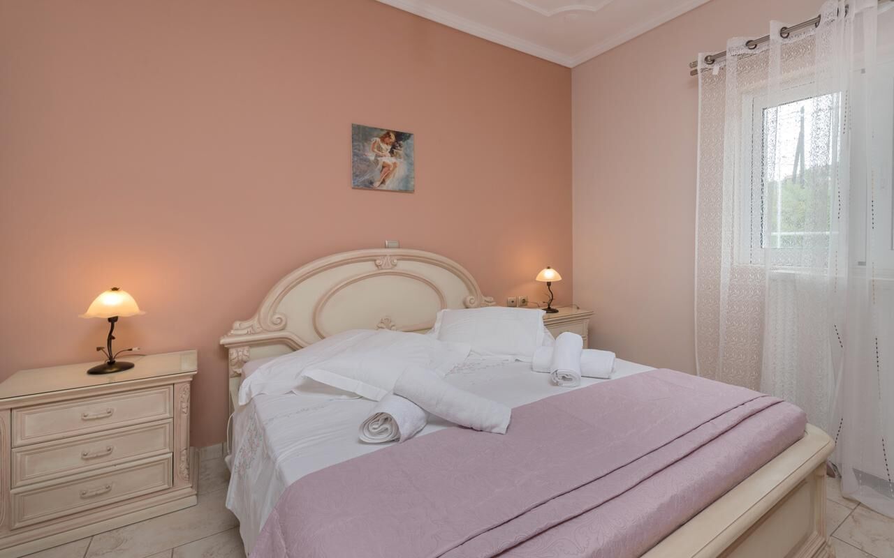 A neatly made bed with white linens and a pink blanket, two bedside lamps, a nightstand, and a painting on the wall.