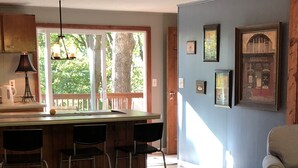 A living room with a floral sofa, a dining area with a bar, and a view of trees through a large window. - Winter mtn views 2 min. to center, fireplace: Monthly rentals welcome! (Columbus)
