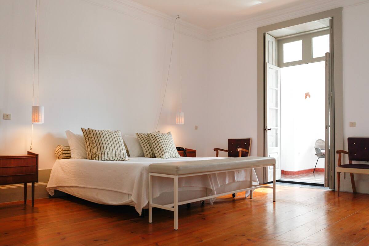 Un dormitorio con cama, mesitas de noche, una silla y una puerta que da a un espacio exterior.